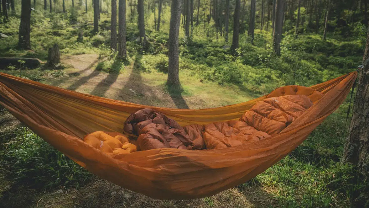 Campingartikel, orange Hängematte mit einem Schlafsack darin zwischen den Bäumen in einem Wald