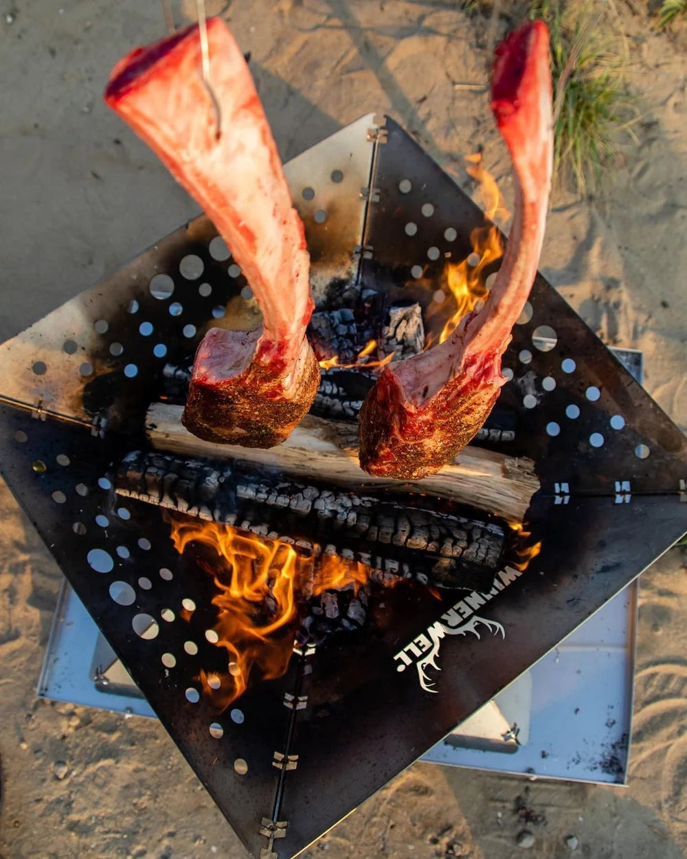 Winnerwell Flache Feuerschale von oben mit zwei Stücken Fleisch am Knochen über dem Feuer auf Sand