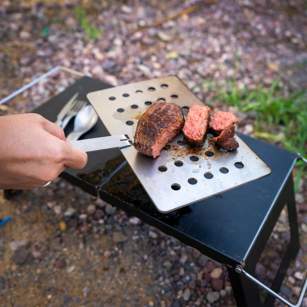 Winnerwell Grillplatte aus Edelstahl mit geschnittenem Steak auf einem Tischchen, eine Hand hält den Stiel