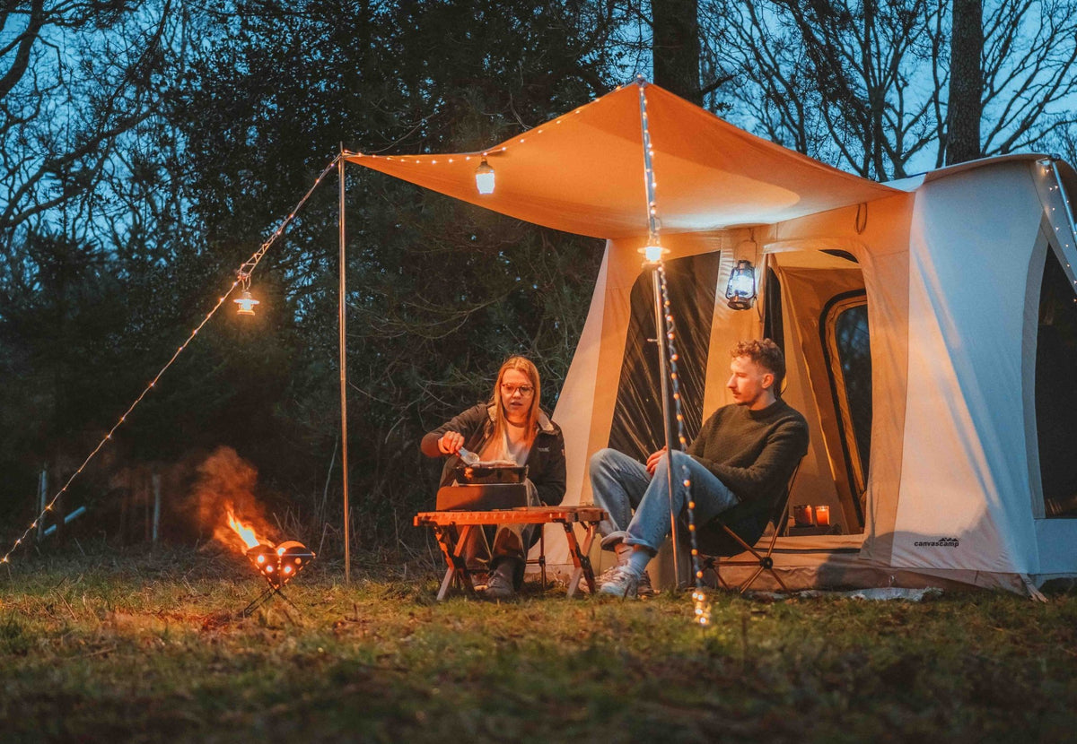 CanvasCamp Big Horn am Abend auf dem Campingplatz mit beleuchtetem Vordach, Lagerfeuer und zwei Campern