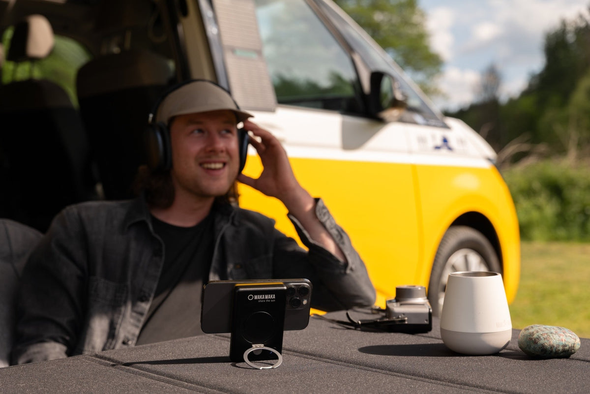 WakaWaka Power Pro mit Handy auf einem Tisch im Freien, ein Mann mit Kopfhörern vor einem gelben Bus