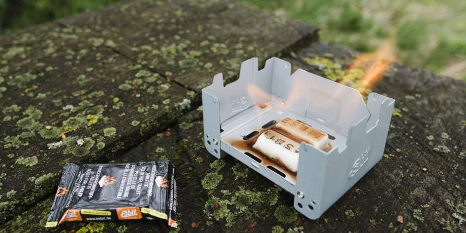 Esbit Survival Stove im Einsatz auf einer bemoosten Steinmauer mit Flammen und einer Packung Tabletten daneben