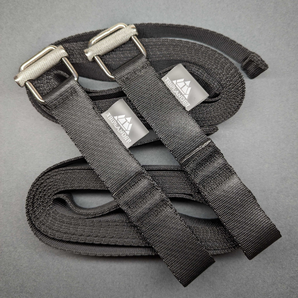 Triplander Quick Drag, zwei schwarze Gurte mit Schnallen und Logoetiketten auf aufgerolltem Gurtband