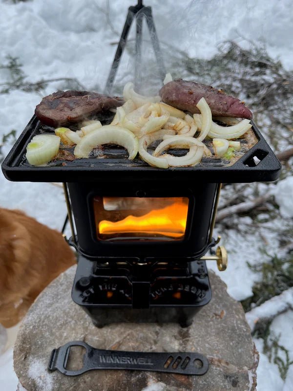 Winnerwell Iron Camping Cooker Stove im Schnee mit Fleisch und Zwiebel auf der Grillplatte und Feuer hinter dem Fenster