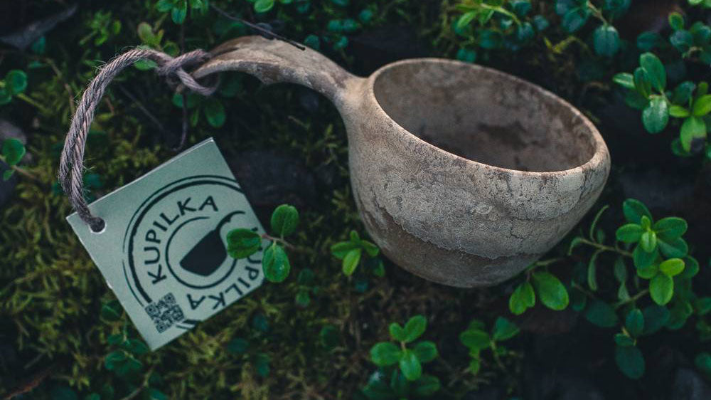 Kupilka, hellgraue Kuksa mit einer Schnur und einem Etikett zwischen grünem Moos