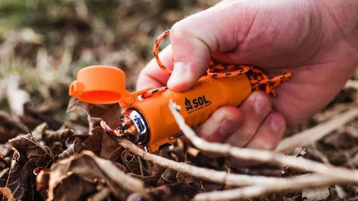 Feuerzeuge und Feuerstarter, Hand mit einem orangen Feuerstarter an einer Schnur über trockenem Laub