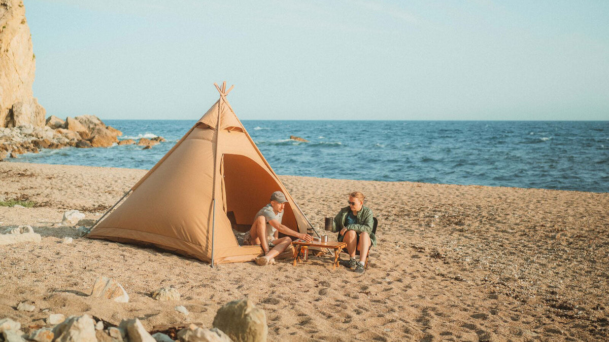 Pyramidenzelte, beiges Pyramidenzelt am Strand mit zwei Personen an einem kleinen Tisch am Meer