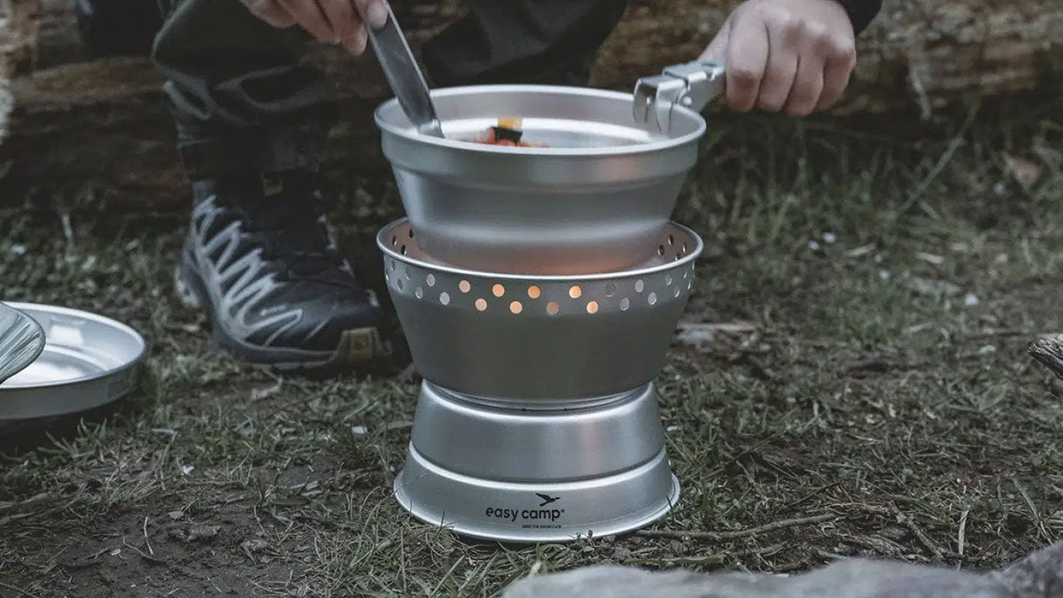 Topfsets, Hand rührt in einem Topf auf einem Campingkocher mit Windschutz draußen auf dem Boden