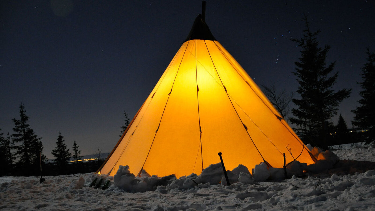 Tipi Zelt, das von innen leuchtet, im Schnee unter einem Sternenhimmel zwischen Kiefern