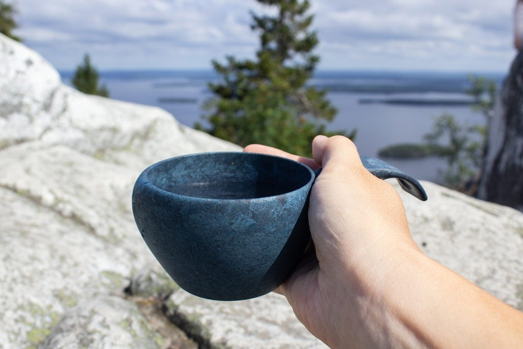 Blaue Kupilka 21 Classic Cup in der Hand auf einem Felsen mit Blick über einen See