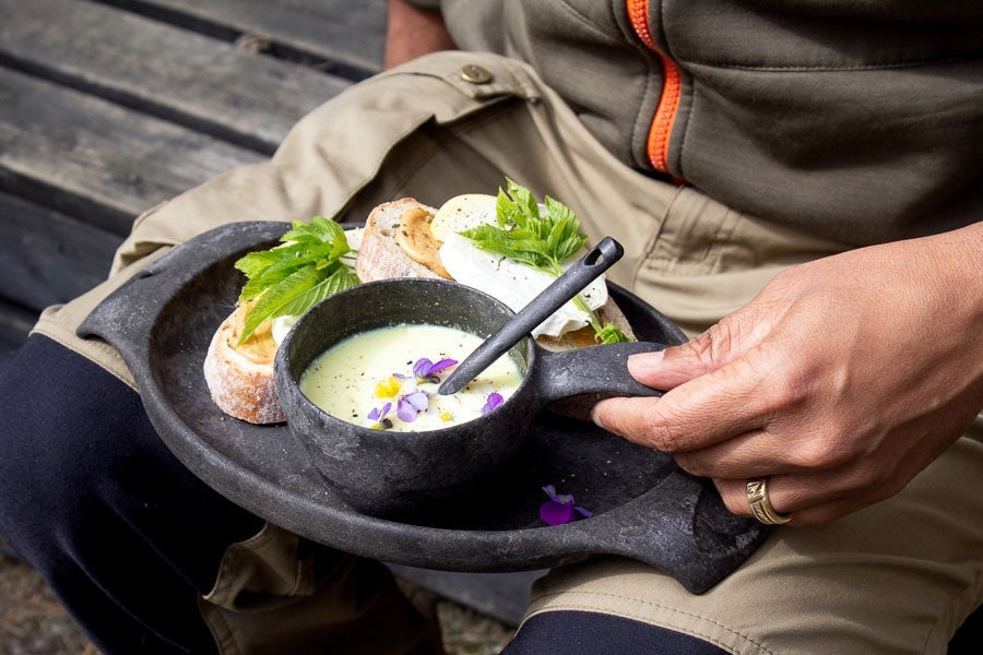 Schwarze Kupilka 21 Classic Cup mit Suppe und Löffel auf schwarzem Teller mit Brötchen, auf dem Schoß