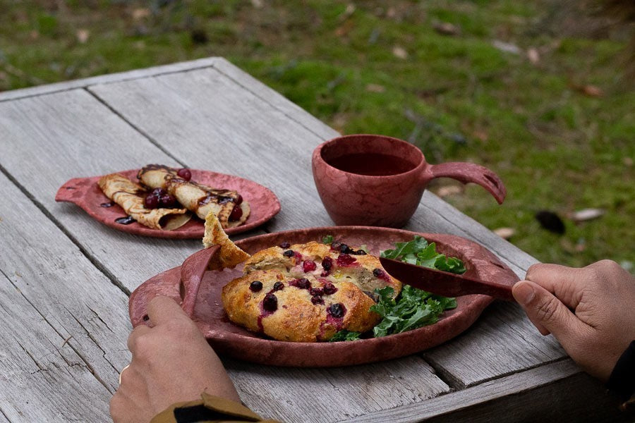 Roter Kupilka 44 Teller mit herzhaftem Gebäck und Salat auf einem Holztisch im Freien, neben einem roten Becher