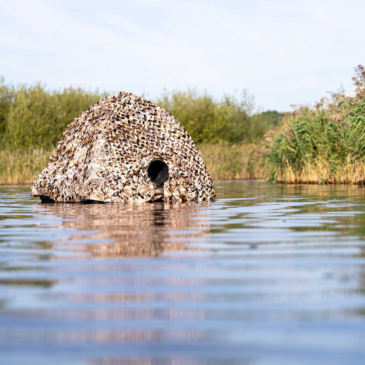 Buteo Tarnnetz 4 über einem Tarnversteck im Wasser, mit Objektivöffnung und Schilf am Ufer