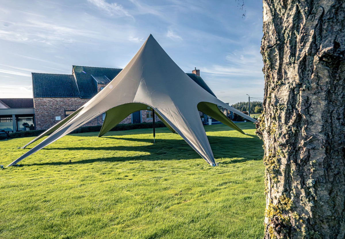 CanvasCamp StarShade Pro Eventzelt in Grau auf einem Rasen vor einem Haus, an einem Baum vorbei gesehen