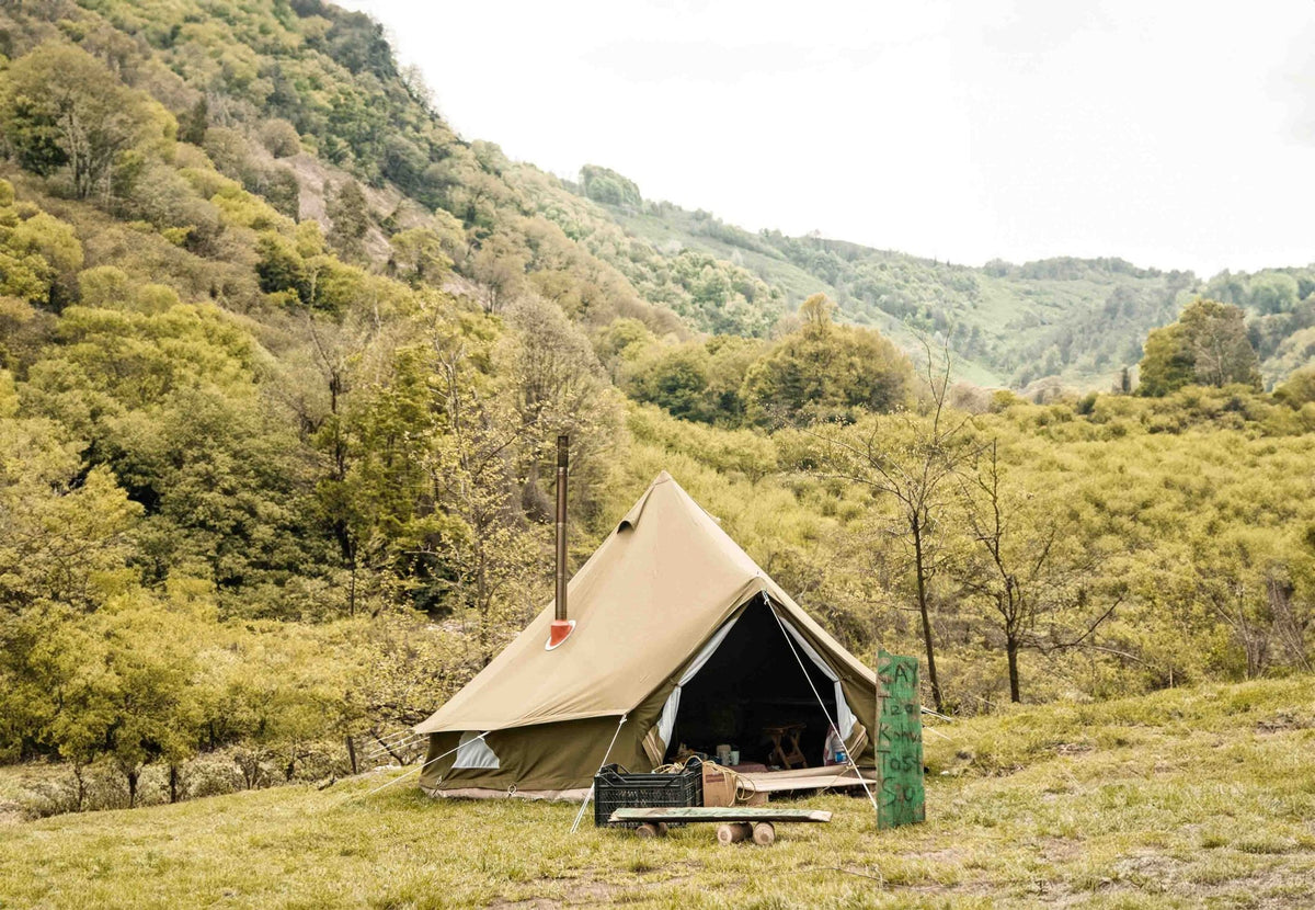 CanvasCamp Exit 45° auf einem beigen Zelt mit offener Tür und Ofenrohr in einer grünen Berglandschaft