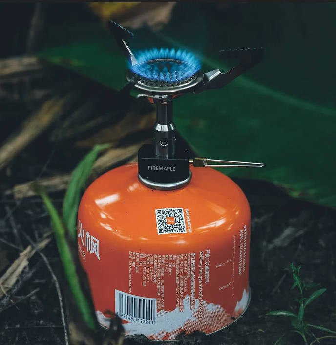 Fire Maple BUZZ auf einer orangefarbenen Gaskartusche mit blauer Flamme, draußen im Gras