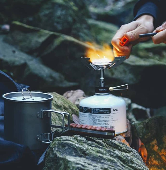 Fire Maple BUZZ auf einer Gaskartusche zwischen Felsen, mit einem Feuerzeug angezündet, daneben ein Topf