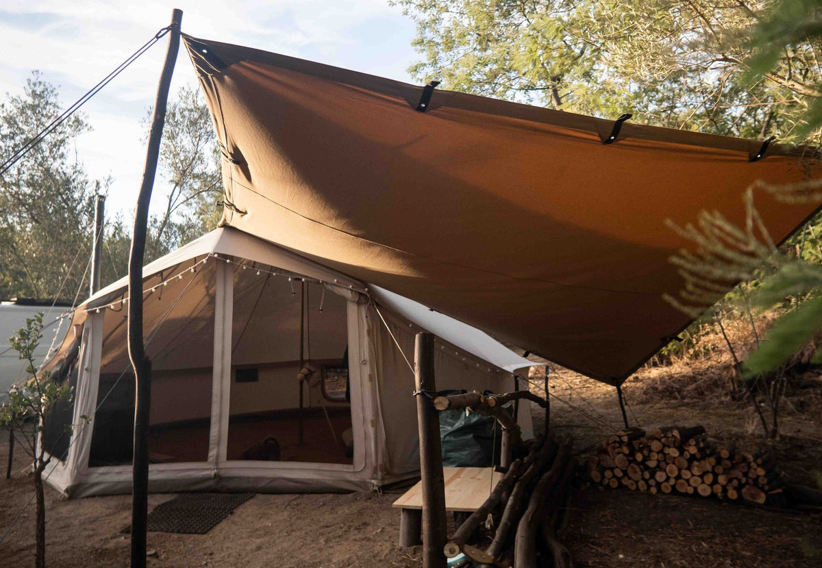CanvasCamp Heavy Duty Shelter in Braun über einem Zelt gespannt, daneben ein Holzstapel