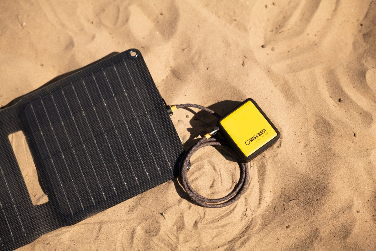 WakaWaka Power 10 im Sand, an ein schwarzes faltbares Solarpanel angeschlossen