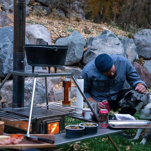 Winnerwell Fire Guard Stove Table an einem brennenden Ofen mit Pfanne darüber und Essen auf dem Rand