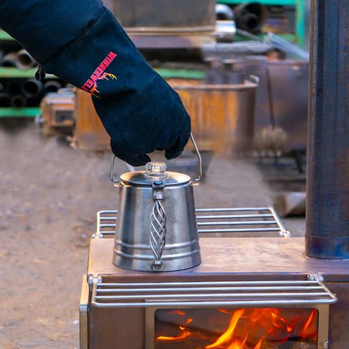 Winnerwell Handschuh hebt einen Metallkessel von einem brennenden Holzofen