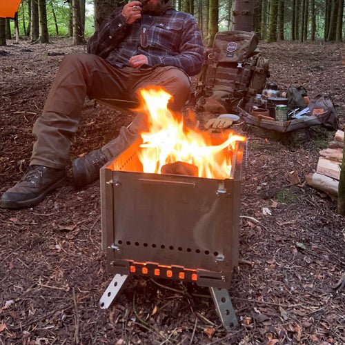 Winnerwell Grill Firepit mit brennendem Holzfeuer im Wald, ein Mann sitzt dahinter